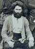 man seated, light colored jacket, full beard, dark hair